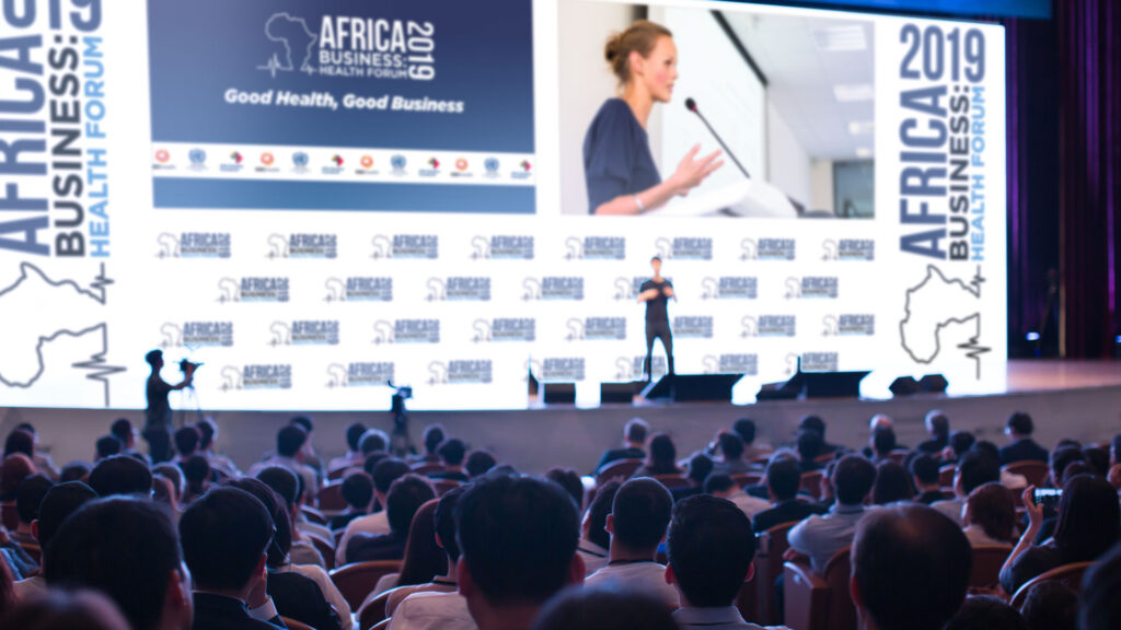 conference stage with speaker and logo backdrop and live tv feed projected